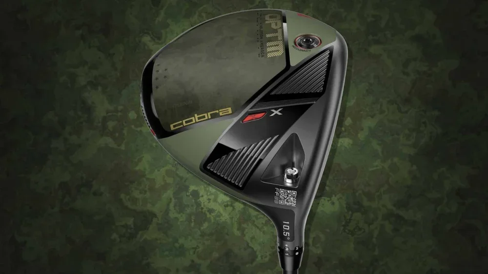 A close-up of a Cobra golf driver with a black and olive green volition design, featuring the brand name and a QR code on the clubhead, set against a camouflage-patterned background.